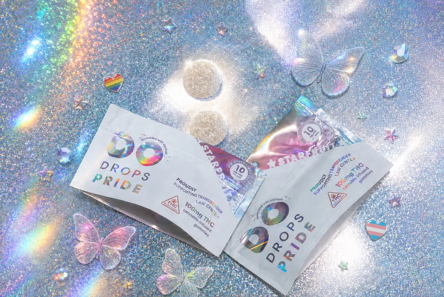 Two DO Drops Pride Starfruit pouches opened on a glittering iridescent surface, with sugared gummies, holographic butterflies and rainbow and transgender pride pins scattered around them.